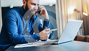 Smiling bearded businessman sitting at table in front of computer, talking on cell phone, showing pen on laptop screen