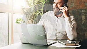 Young smiling bearded businessman is sitting at table in front of laptop, drinking coffee, talking on cell phone.