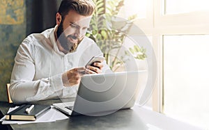 Young smiling bearded businessman sitting at table in front of computer, using smartphone. Man checking email, chatting.