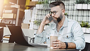 Young smiling bearded man having mobile phone conversation with business partner, while sitting with laptop in cafe