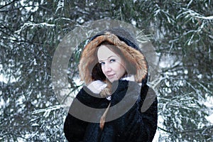 Young smiling beautiful woman with blue eyes in black faux fur coat with red hood on background of winter snowy park