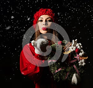 Young smiling beautiful woman dressed in red winter sweater, hat and mittens holding Christmas ornate wreath and sending air kiss