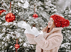 Young smiling beautiful woman in fur coat and mittens standing near decorated Christmas tree and catching snowflakes