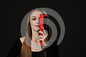 Young smiling beautiful woman with long silky straight hair in black clothes holding red hair straightener in hand over