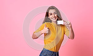 Young smiling beautiful woman in yellow shirt showing credit card in hand over pink background