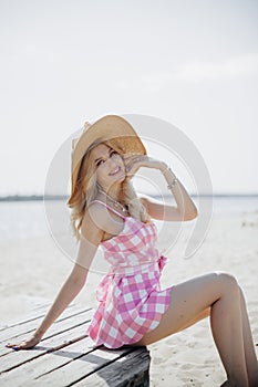 Young smiling blonde girl look like a Barbie doll in pink mini dress and wide brim hat sitting on beach