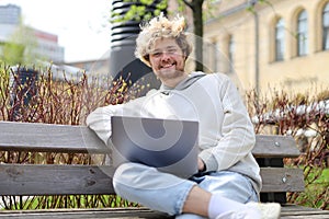 A young smiling blonde sits with a laptop on a bench in a city park