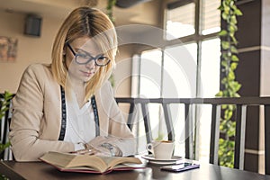 Young smiling blonde woman in a restaurant reading a book and drinking coffee