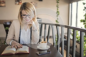 Young smiling blonde woman in a restaurant reading a book and drinking coffee