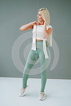 Young smiling blonde woman in sportswear doing stretching on background of grey wall