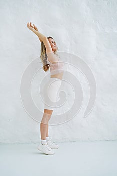 Young smiling blonde woman in sportswear doing stretching on background of white wall