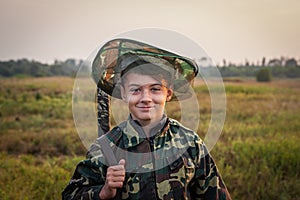 Young smiling boy with hunting shotgun standing at green field during sunset