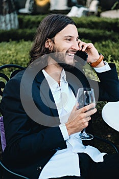 Young smiling brunette bearded businessman with glass of red wine happily talking on cellphone in restaurant outdoor