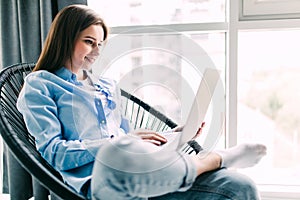 Young smiling brunette girl is sitting on modern chair near the window in light cozy room at home working on laptop in relaxing