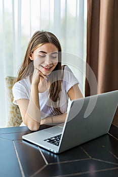 Young smiling girl is sitting on modern chair near the window in light cozy room at home working on laptop in relaxing atmosphere