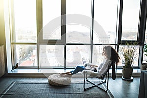 Young smiling brunette girl is sitting on modern chair near the window in light cozy room at home working on laptop in relaxing
