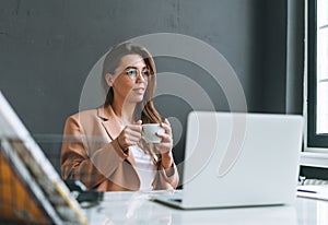 Young smiling brunette woman in glasses with long hair in beige suit resting and drinking coffee on working place in the modern