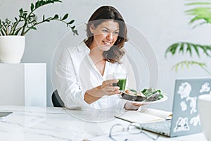 Young smiling brunette woman nutritionist doctor plus size in white shirt with green healthy food at modern bright office room. photo