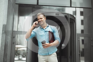 Young smiling business man with cup of coffee and laptop in his hands talking on the phone standing near entrance to