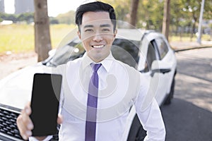 Young smiling business man showing mobile phone with black empty screen and standing before the car