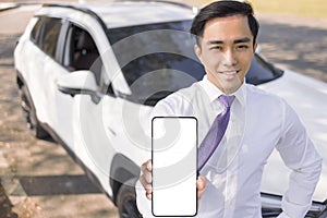 Young smiling business man showing mobile phone with white empty screen and standing before the car