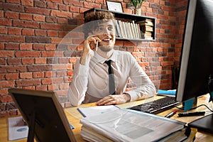 Young smiling business man talking by smartphone at working place at computer