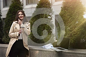 Young smiling businesslady having pleasant phone conversation while working in her laptop and drinking coffee , stands in the