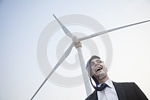 Young smiling businessman standing beside a wind turbine, tie is on his head