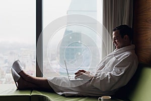 Young smiling businessman working on laptop computer wearing white bath robe sitting near window with cup of coffee