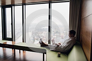 Young smiling businessman working on laptop computer wearing white bath robe sitting near window with cup of coffee