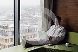 Young smiling businessman working on laptop computer wearing white bath robe sitting near window with cup of coffee