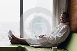 Young smiling businessman working on laptop computer wearing white bath robe sitting near window with cup of coffee