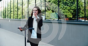 Young smiling businesswoman in a business suit while riding an electric scooter drinking coffee on the background of the