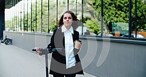 Young smiling businesswoman in a business suit while riding an electric scooter drinking coffee on the background of the