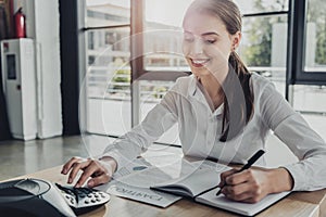 young smiling businesswoman doing paperwork and pushing button of conference phone on table