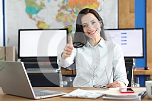 Young smiling businesswoman showing thumbs up gesture while sitting at her working table in office