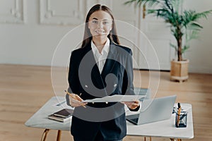 Young smiling bussiness woman looking at camera and holding document in front of office desk