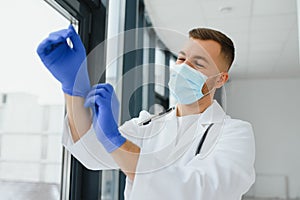 Young smiling Caucasian male doctor in white uniform, with eyeglasses and stethoscope around neck putting rubber gloves before