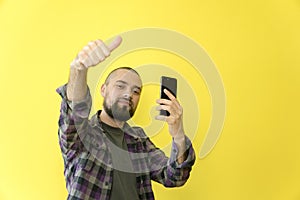 Young smiling caucasian man with a beard and a bald head holding a phone in his hands on a yellow background. Approval, thumbs up