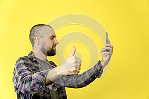 Young smiling caucasian man with a beard and a bald head holding a phone in his hands on a yellow background. The blogger