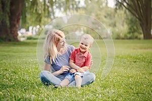 Young smiling Caucasian mother and laughing boy toddler son sitting on grass in park. Family mom and child hugging having fun