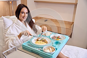 Young smiling caucasian pregnant woman wearing bathrobe cutting meat