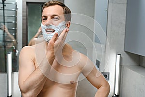 Young smiling Caucasian shirtless man applying shaving foam on face in front of mirror, preparing for hair removal
