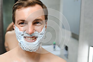 Young smiling Caucasian shirtless man applying shaving foam on face in front of mirror, preparing for hair removal