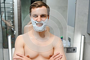 Young smiling Caucasian shirtless man applying shaving foam on face in front of mirror, preparing for hair removal