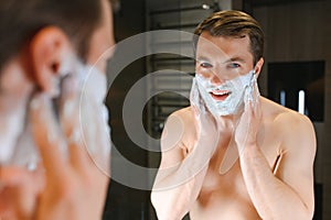 Young smiling Caucasian shirtless man applying shaving foam on face in front of mirror, preparing for hair removal