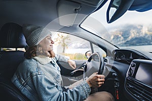 Young smiling caucasian woman driver traveler in a hat and jacket driving a car while traveling along the misty mountains