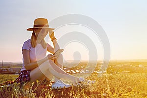 Young smiling caucasian woman in a hat uses a mobile phone sitting on the grass in a park at sunset. Happy girl writes a message