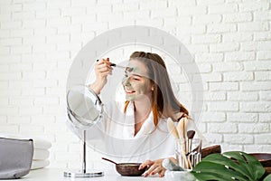 Young smiling caucasian woman wearing bathrobes appplying clay face mask looking at the mirror