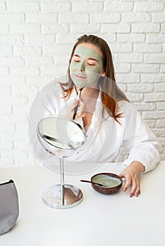 Young smiling caucasian woman wearing bathrobes appplying clay face mask looking at the mirror